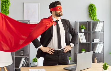 Portrait of confident young funny man in superhero face mask and red cloak standing at the desk on...