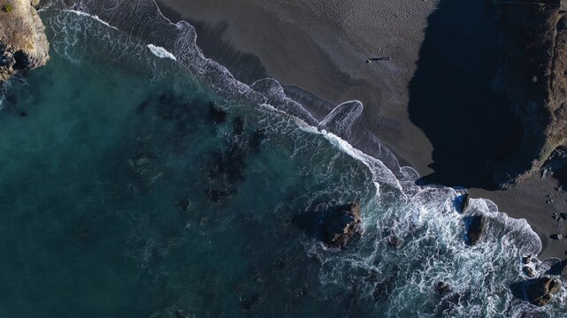 Aerial view of turquoise waters crashing against a dark sandy beach under the warm sun, coastal bluffs framing the scene, Jenner, California, United States.