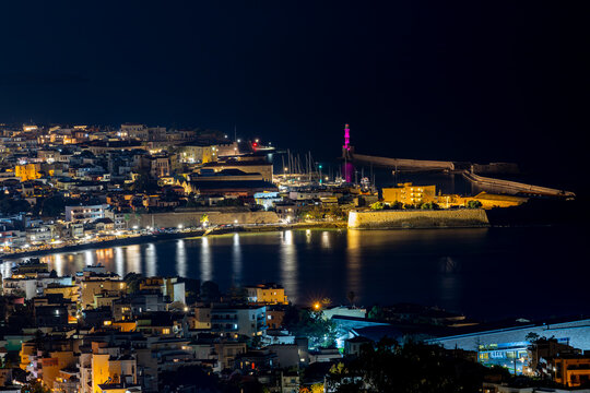 Aerial view of the illuminated Venetian Lighthouse standing tall against the inky sky, casting a vibrant glow over the serene harbor waters, Chania, Chania, Greece.