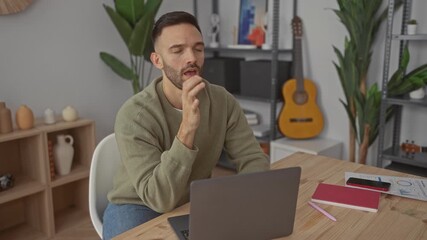Young hispanic man sitting in a living room at home using a laptop appears frustrated with paperwork scattered on a table surrounded by indoor plants and guitar decor - Powered by Adobe