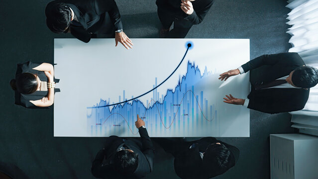 A group of business professionals collaborates during a meeting, analyzing financial data on a digital graph displayed on a table to strategize their next moves. Scalp