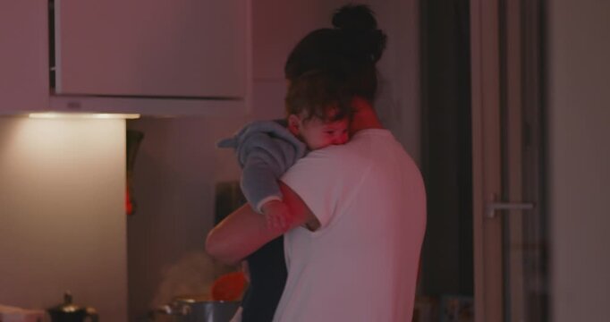 Mother stirring pot while holding baby in arms, balancing caregiving and cooking during evening family routine at home
