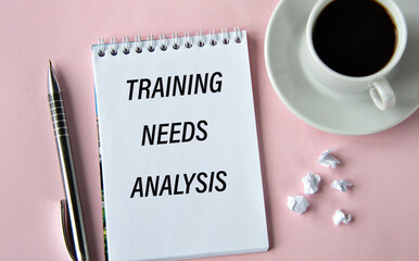 TRAINING NEEDS ANALYSIS - words in a notebook on a pink background with a cup of coffee