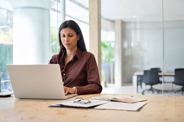 Young professional it specialist latin hispanic business lady typing on laptop pc sitting at desk in office. 30s middle eastern indian woman using computer technology app for work online. Copy space