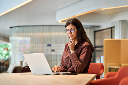 Young professional it specialist latin hispanic business lady working on laptop pc sitting at desk in modern office space. Focused middle eastern indian woman using computer technology for work online