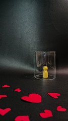A small yellow figurine stands trapped under a clear glass cup while red heart shapes lie freely around it, symbolizing isolation, emotional distance, and unreachability in love on a dark background.