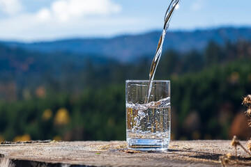 Stream of fresh water pouring into glass against blurred forest and mountain background, eco lifestyle. © Oleksandra