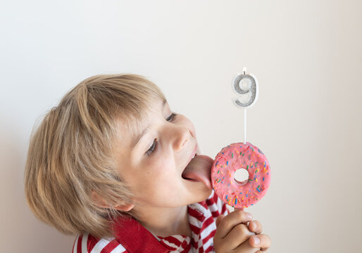 9-year-old boy holds donut on a stick with burning candle in shape of number nine and licks it with his tongue. A contented child, a sweet life, a birthday party. Positive emotions, a happy childhood