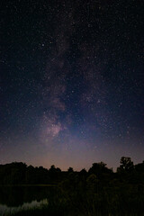 Fototapeta premium Stunning night landscape with the Milky Way stretching across the starry sky above a calm lake and dark forest silhouettes