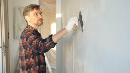 Male construction worker wearing protective gloves smoothing plaster onto wall surface during home...