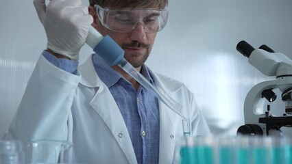 Scientist man wearing a lab coat and safety glasses is using a micropipette to drop a liquid into a...