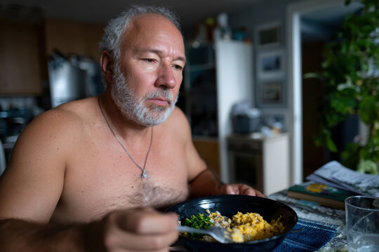 A middle-aged man sits shirtless at a table savoring a hearty breakfast of eggs and greens, capturing a moment of personal time and nourishment in a cozy kitchen. - Powered by Adobe