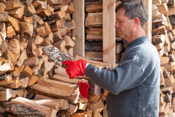 A man in red gloves picking up firewood from a large pile. An adult man in a gray sweater picks up firewood, against the backdrop of a large, neatly stacked woodpile