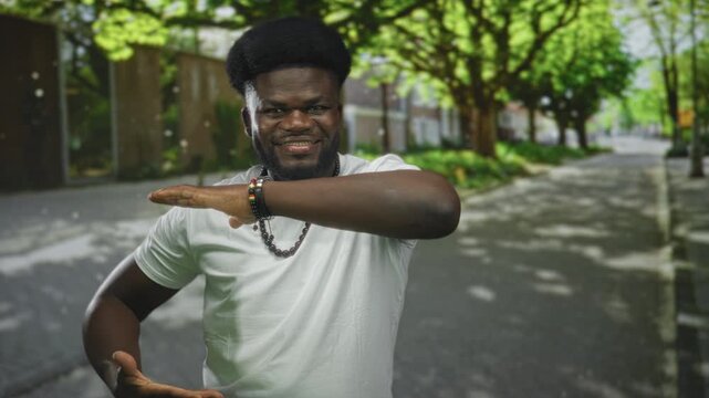 Man dabbing with arm across face on sunny street wearing white t shirt and beaded necklace; playful confidence joy.