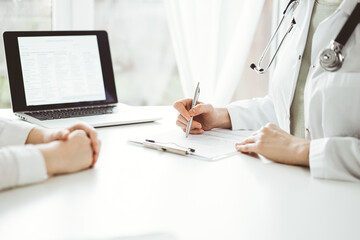 Doctor and patient sitting near each other at the white desk in clinic. Female physician is listening filling up a records form. Medicine concept