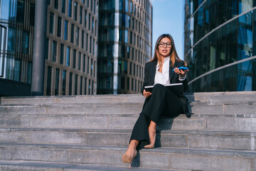 Businesswoman in a suit sits on steps speaking on her phone, surrounded by impressive urban architecture.