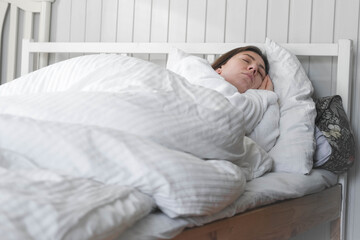 A woman is sleeping peacefully in her bed with a white pillow and a blanket. A beautiful young happy lady is sleeping peacefully on a cozy bed in the morning light at home, in a relaxed atmosphere