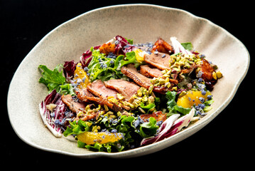 A gourmet duck breast salad with mixed greens, roasted sweet potatoes, and orange segments, garnished with chopped pistachios and edible flowers in a shallow bowl on a black background