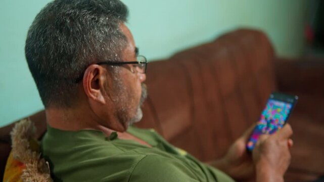 A side profile close-up of a middle-aged Asian man with a beard and glasses, sitting on a sofa and intently playing a mobile game on his smartphone.