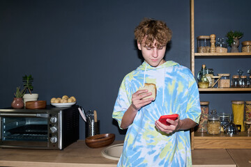 Teenage boy standing in kitchen holding sandwich in one hand and using smartphone with other hand, looking at device screen while preparing to eat snack © Mediaphotos