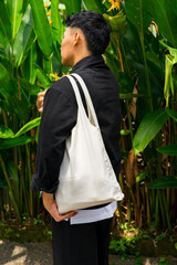 A man wearing black shirt and white tote bag standing in front of tropical plants, showing sustainable lifestyle and eco-friendly fashion concept.