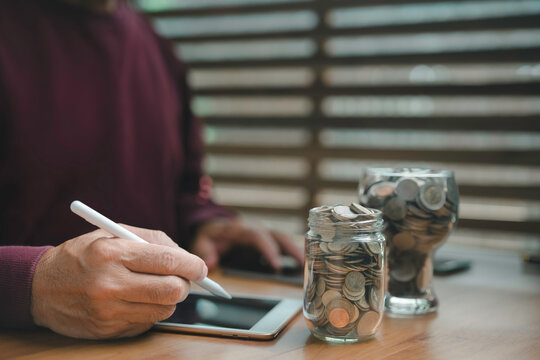 Close-up of a person using a digital tablet with a stylus beside glass jars filled with coins, symbolizing financial planning, online banking, saving money, investment, and modern technology.