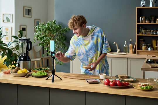 Young boy preparing sandwich while recording video on smartphone in modern kitchen, holding bread and interacting with device, fresh vegetables and blender on countertop