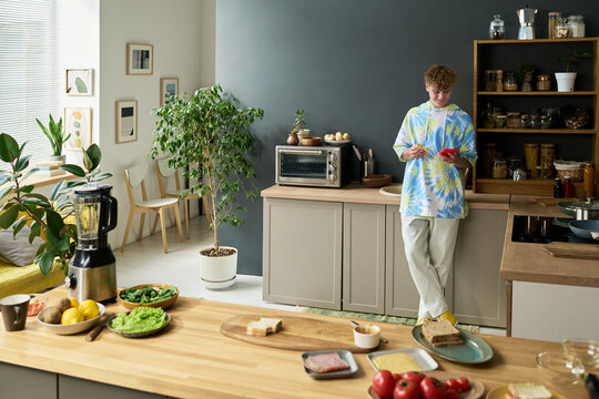 Teenage boy standing in modern kitchen using digital tablet, surrounded by fresh vegetables and kitchen appliances, preparing food while leaning against counter