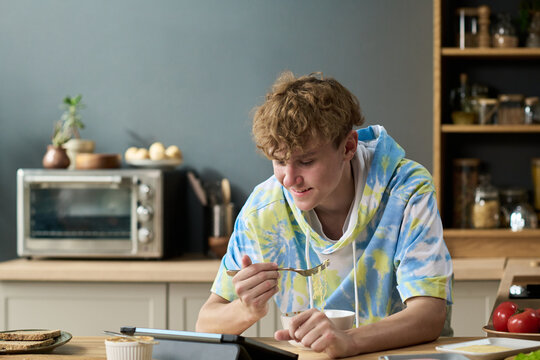 Teenage boy sitting at kitchen counter eating breakfast while looking at digital tablet, smiling and engaging with screen, kitchen appliances and food in background
