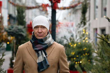 Winter portrait of serious caucasian man wearing white beanie and cozy scarf at Christmas festive decorated modern city. Cold winter weather.