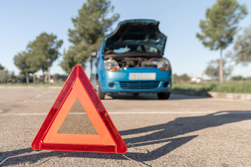 Red warning triangle placed on asphalt near a broken car with open hood, signaling vehicle breakdown