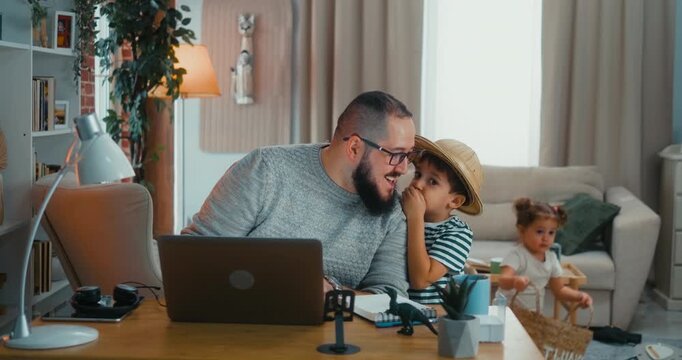 Little boy approaches his father and whispers something while he works on the computer. Sweet moment of affection interrupting focus in a real everyday home scene.