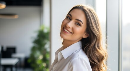 Obraz premium Smiling Young Woman in White Shirt Posing in Modern Office with Natural Light, Exuding Confidence and Professionalism