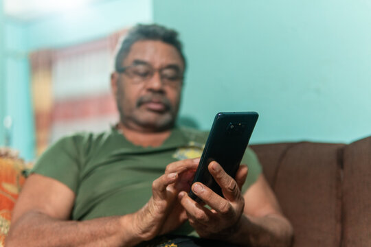 A side profile close-up of a middle-aged Asian man with a beard and glasses, sitting on a sofa and intently playing a mobile game on his smartphone.