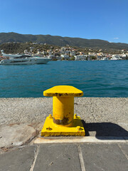 Bright yellow metal bollard casts a strong shadow at the harbour edge against vivid blue sea and sky.