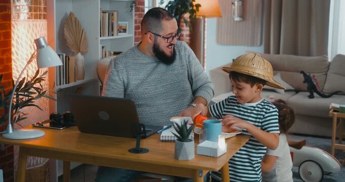 Playful moment as kids interrupt their father working online, bringing toys and pretend drinks. He joins their game while still at his desk, showing fun balance of remote work and parenting.