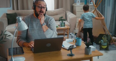 Father having an online video conference call at his home desk while kids play in the background with a toy tent. Real work-life balance and multitasking parenting in a modern cozy home.