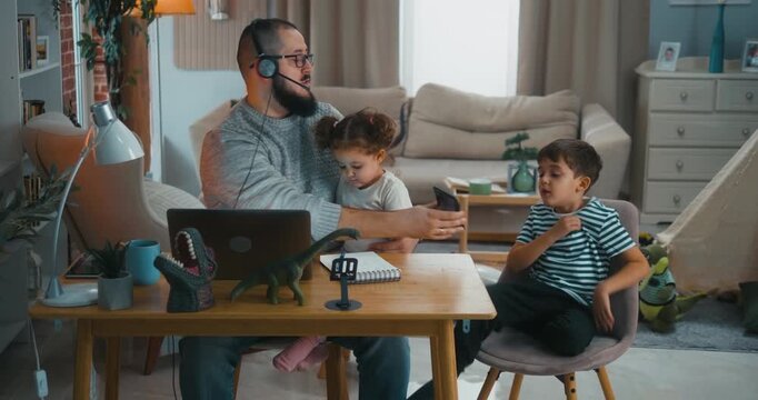 Father gives smartphones to his kids and starts remote work on his laptop, showing modern reality of digital distraction and screen dependence in everyday family life at home.