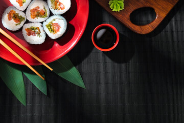 A red plate with maki rolls and banana leaves, chopsticks, a bowl of soy sauce, wasabi, and a cutting board on a black bamboo mat in soft sunlight.