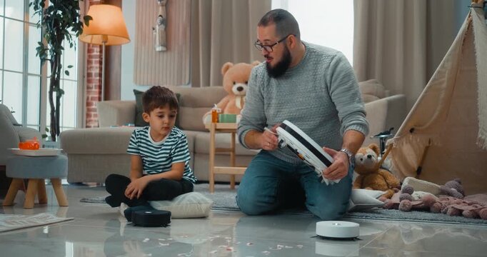 Father teaches his son how a smart robot vacuum works, showing features and explaining technology at home. Positive parenting, learning and modern family life with helpful smart devices.
