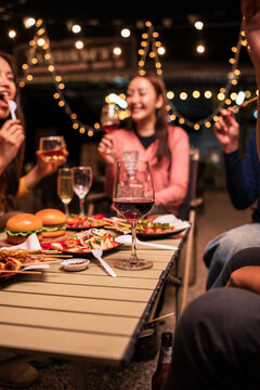 A group of cheerful friends enjoying a night out at an outdoor restaurant. They are seen toasting with various drinks including wine, beer, and whiskey over a table filled with delicious food like bur