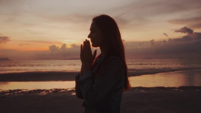 Silhouette of woman with folded hands praying against beautiful sunset near sea concept of humility religion and meditation. Believer woman praying to God