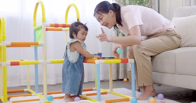 Asian mother smiling beside toddler girl playing with colorful blocks near sofa, encouraging curiosity and creativity, concept of home education, family happiness, and early child learning