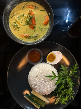 A bowl of shrimp in spicy coconut milk (udang lemak cili api) served alongside rice, sambal, boiled egg, cucumber, and ulam on a dark plate, showcasing vibrant Malaysian flavors. Top view.