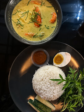 A bowl of shrimp in spicy coconut milk (udang lemak cili api) served alongside rice, sambal, boiled egg, cucumber, and ulam on a dark plate, showcasing vibrant Malaysian flavors. Top view.