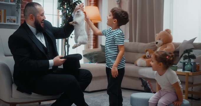 Father dressed as a magician surprises his excited children by pulling a white rabbit from a hat during a fun home magic show. Joy, laughter and creative family entertainment in the living room.
