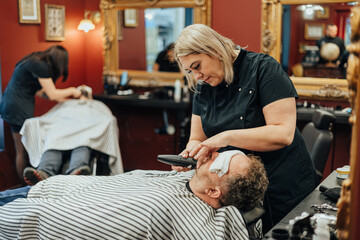 Two barbers trim beards of clients lying back with eye towels in a modern barbershop with red walls and ornate mirrors