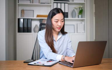 Focused Asian businesswoman working on a laptop at a desk in a modern office environment.