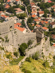 Obraz premium Ancient stone walls of Kotor's fortress snake through lush Montenegrin hills, overlooking a charming village of red-tiled roofs and winding roads.