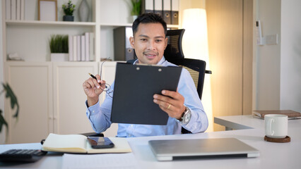 Confident professional reviews documents on a clipboard at a bright home office, pen in hand and glasses lifted.